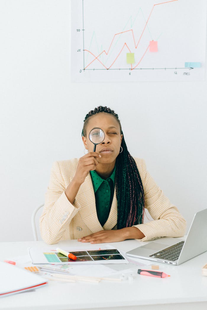 Woman examining data charts with a magnifying glass at a desk with a laptop, showcasing analytical skills.